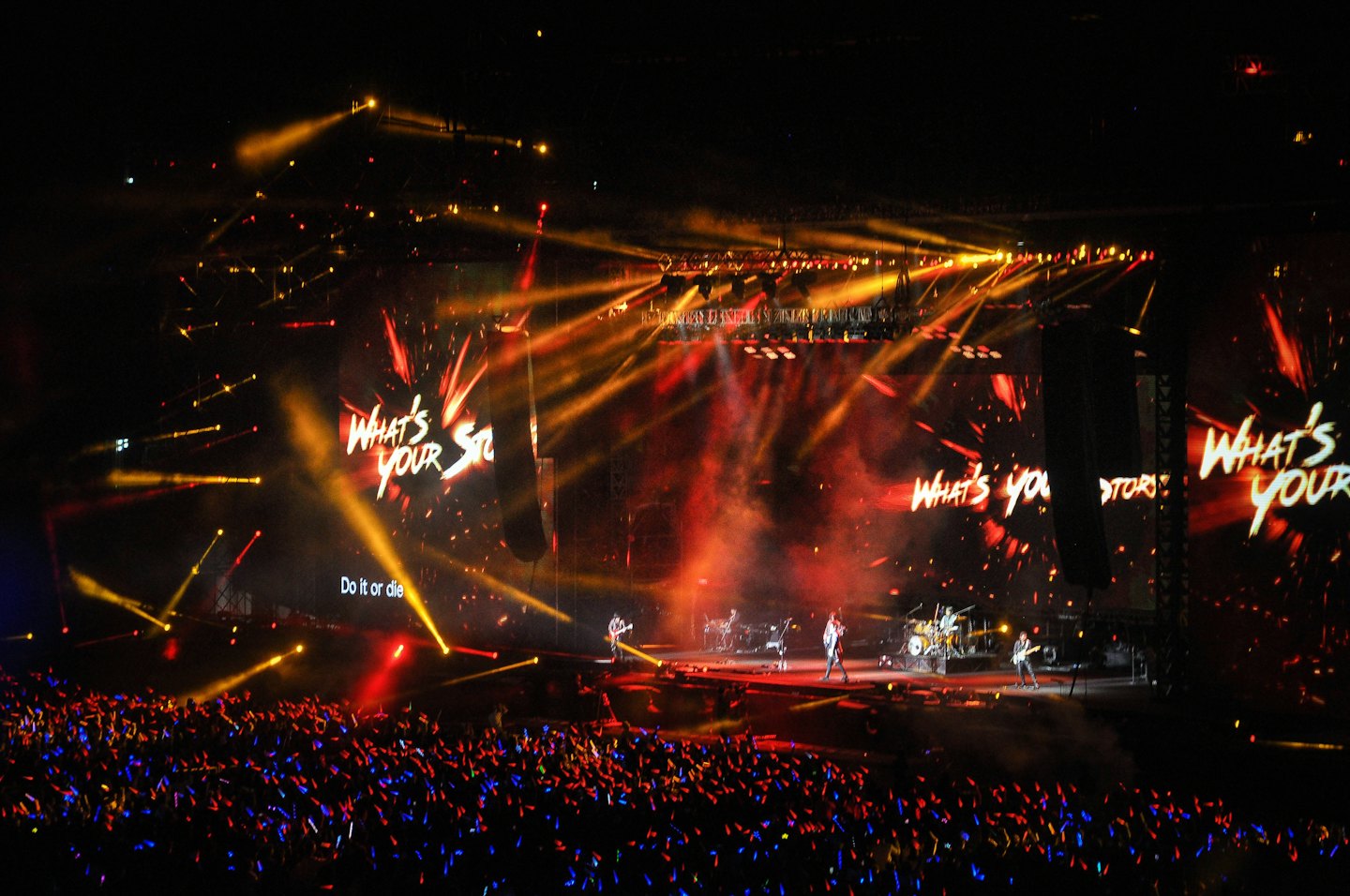 A band performs on stage before a crowd waving glowing sticks.